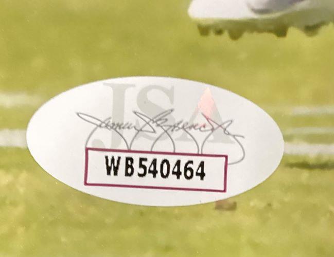 A Sports Integrity oval sticker with serial number WB540464, JSA letters, and faint signatures is on the grass next to a Jeremiah Trotter Jr Signed 8x10 Philadelphia Eagles Blocking Photo.
