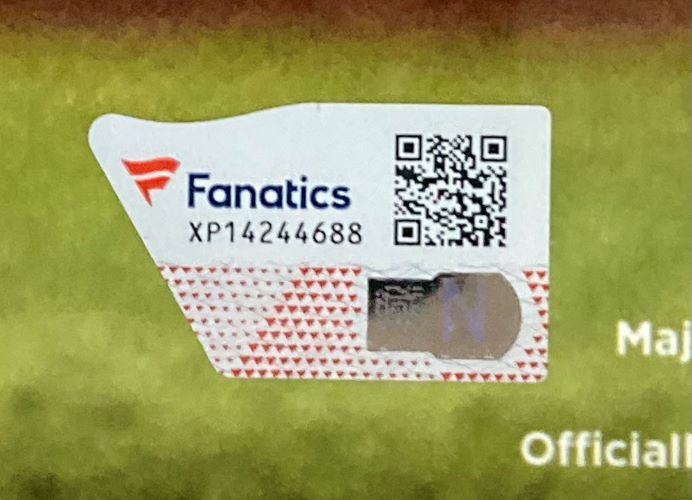 A close-up of a white Fanatics authentication sticker with QR code and serial XP14244688, on an Aaron Judge Signed Framed 16x20 New York Yankees Home Run 62 Photo by Sports Integrity, set against a blurry green and yellow background.