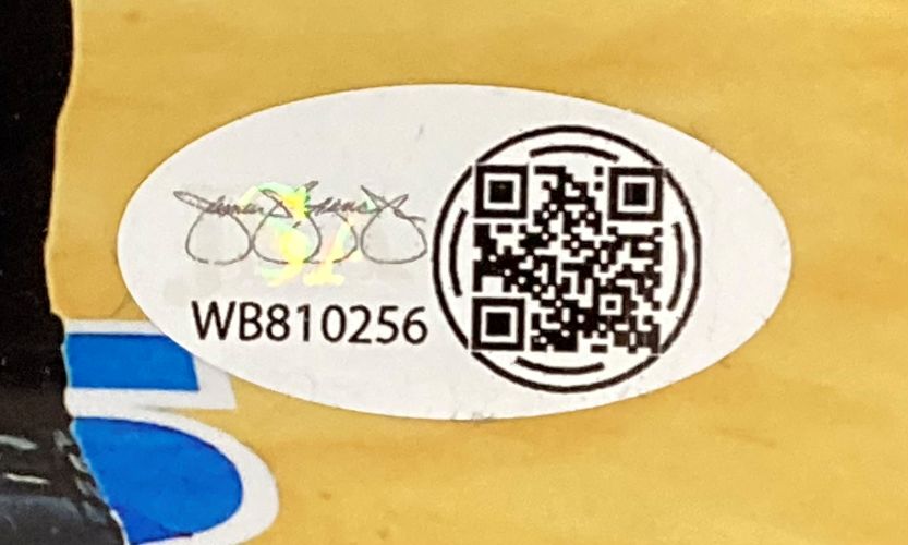 A white oval JSA QR hologram sticker with serial number WB810256 and a faint signature appears on the Amanda Nunes Signed Framed 8x10 UFC Championship Photo by Sports Integrity, placed on a yellow surface with blue at the bottom left.