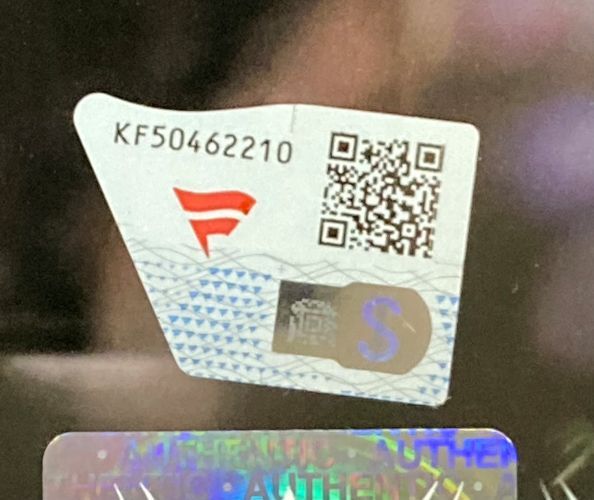 A close-up of a security sticker on the Sports Integrity Bayley Signed Framed 8x10 WWE Wrestlemania XL Photo Fanatics displays code KF50462210, a QR code, a red logo, blue S, holographic features, and partial "AUTHENTIC" text.