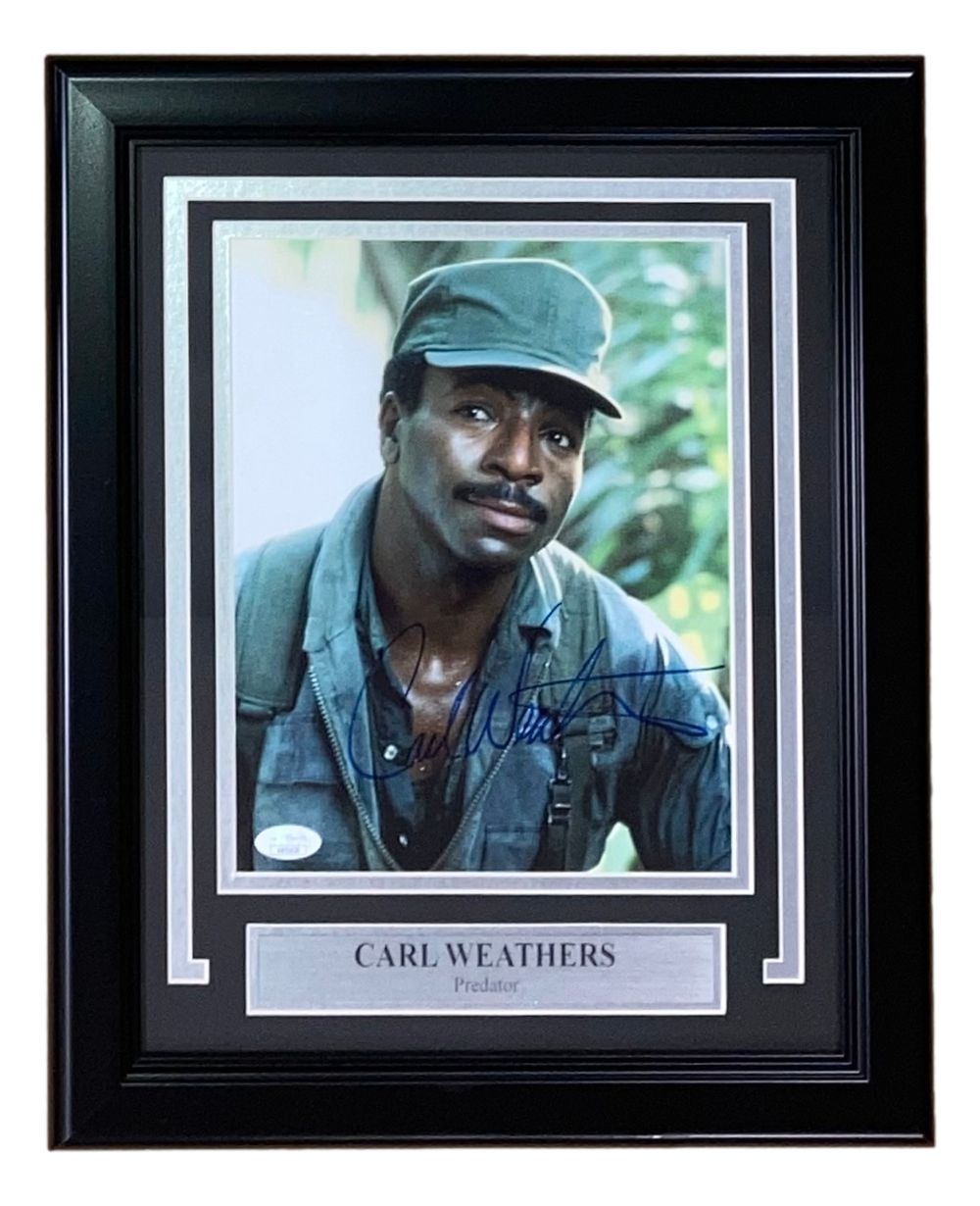 The Sports Integrity Carl Weathers Signed Framed 8x10 Predator Photo (JSA) features a smiling Carl Weathers in military attire, with his autograph and plaque displayed below.