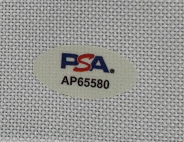 A close-up of the PSA/DNA hologram (code AP65580) on the Chevy Chase Signed Bush Wood Caddyshack Golf Flag by Sports Integrity, shown on a textured white surface.