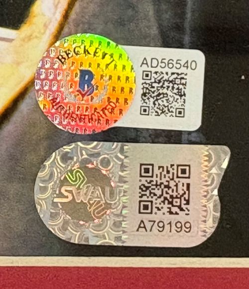 Two authentication stickers with holographic designs, QR codes, and serial numbers (AD56540 and A79199) are affixed next to each other on the Sports Integrity Chris Evans Signed Framed 16x20 Captain America Kneel Photo 2 BAS LOA.
