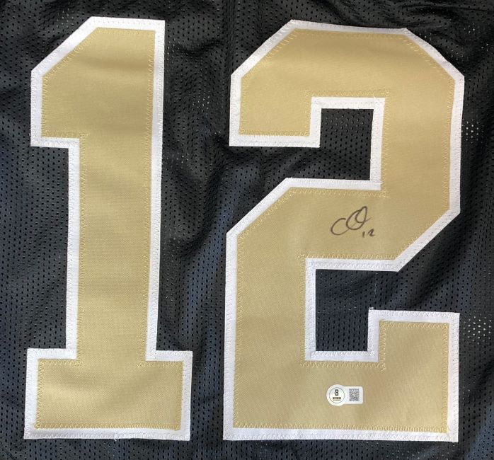 The Sports Integrity Chris Olave New Orleans Signed Black Football Jersey features large gold and white number 12, with Olave's signature in black marker on the "2" and a BAS authentication sticker below it.