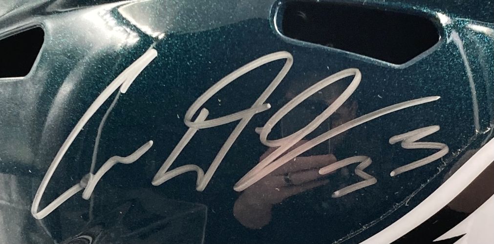 A close-up of the Cooper DeJean Signed Eagles FS Replica Helmet by Sports Integrity, with a silver autograph and "Super Bowl 59 LIX" inscription visible. The shiny surface shows a reflection of someone holding a phone.