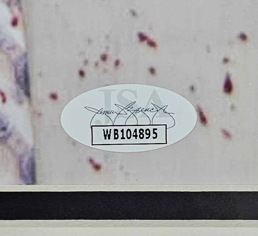A close-up of an oval JSA sticker marked with code WB104895 appears on a Creed Bratton Signed Framed 11x14 The Office Creed Bloody Shirt Photo, authenticated by Sports Integrity, against a light background with blurred spots.