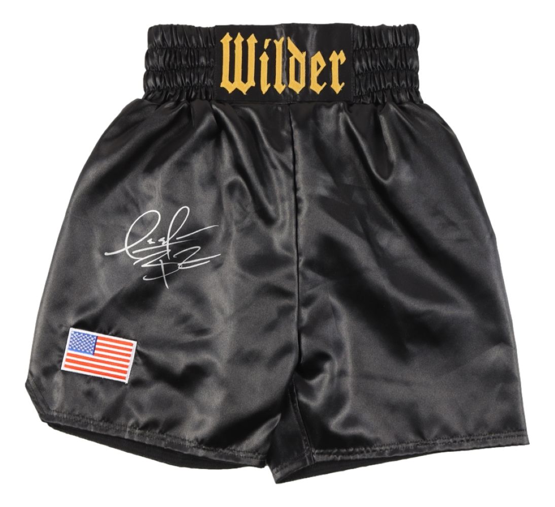 The Sports Integrity Deontay Wilder Signed Custom Black Boxing Trunks feature "Wilder" in gold on the waistband, an American flag patch on the lower left leg, and Wilder’s white signature next to the flag on the left thigh.