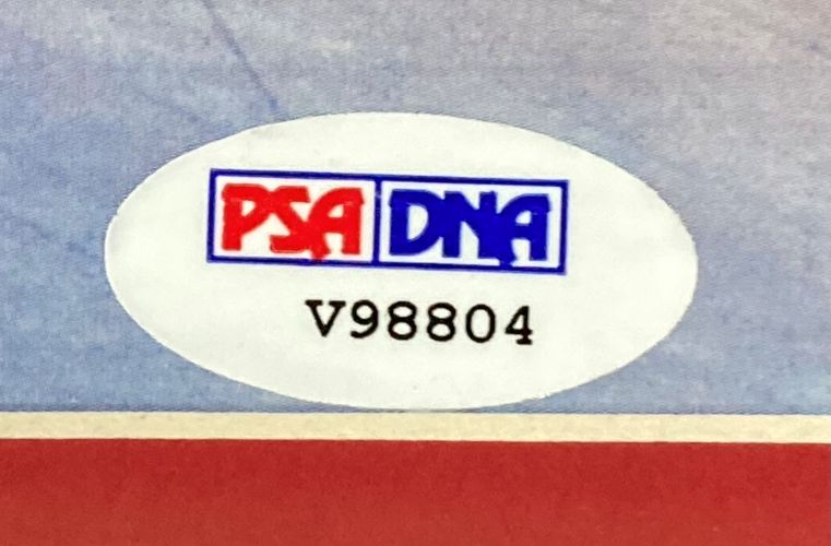 A close-up of a white oval PSA/DNA authentication sticker with a red and blue logo and code V98804 on an Edgar Mitchell Signed Framed 16x20 NASA Apollo 14 Photo by Sports Integrity.