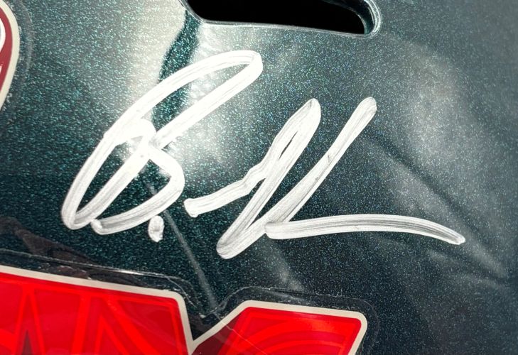 A close-up of the Brandon Graham Signed Eagles FS Super Bowl 59 LIX Replica Helmet by Sports Integrity shows a large silver autograph on its glossy, dark green surface, with part of a red and white sticker at the bottom.