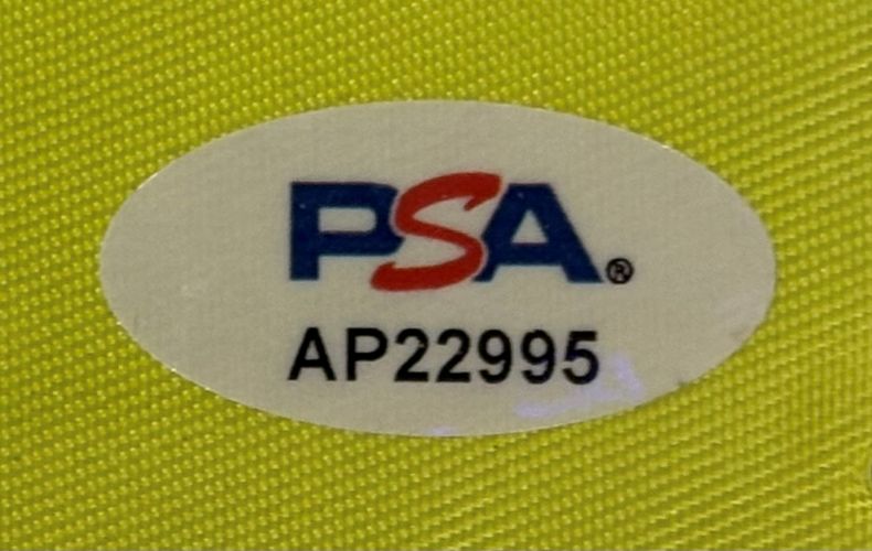 A white oval PSA/DNA hologram sticker (code AP22995) is attached to the Jordan Spieth Signed Framed The Memorial Tournament Golf Flag from Sports Integrity, shown on a textured yellow surface.
