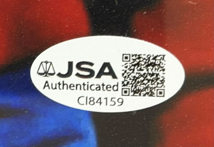 Close-up of a white JSA Authenticated sticker with code CI84159 on a Henry Thomas Matthew De Merritt Signed Framed 11x14 ET Photo by Sports Integrity, set against a blurred red, black, and blue background.