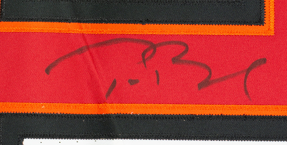 A close-up of Tom Brady's bold, cursive autograph in black marker on a red Tampa Bay Buccaneers Nike Elite Football Jersey from Sports Integrity, bordered with orange and black stitching. Fanatics 975.