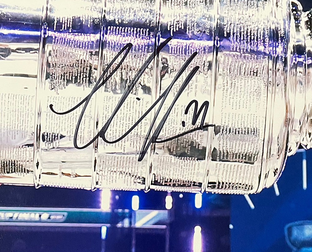 A Victor Hedman signed 16x20 Tampa Bay Lightning trophy photo by Sports Integrity shows a shiny silver trophy with engraved names and a bold black autograph, set against a backdrop of blurred blue and purple lights.
