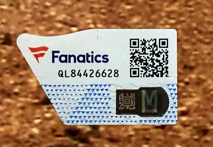 A close-up of a Fanatics authentication sticker with the logo, QR code, and code QL84426628 on a Zack Wheeler Signed Framed 16x20 Philadelphia Phillies Photo by Sports Integrity, set against a blurred brown surface.