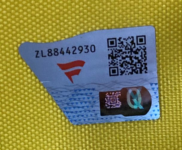 A close-up of a Sports Integrity security label with code ZL88442930, red logo, QR code, and holographic seal on the 2012 Open Championship PGA Issued Golf Flag Fanatics, placed on a textured yellow surface.