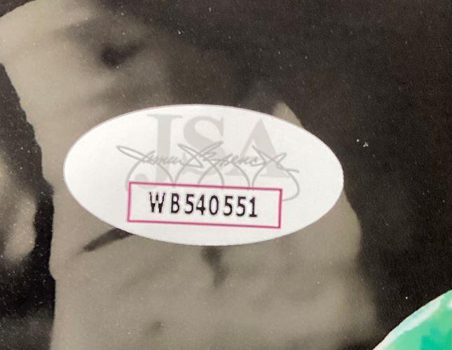 A close-up of a white oval WB540551 sticker appears on a Jeremiah Trotter Jr Signed 11x14 Philadelphia Eagles Spotlight Photo by Sports Integrity, with faint text and signatures in the blurred background.