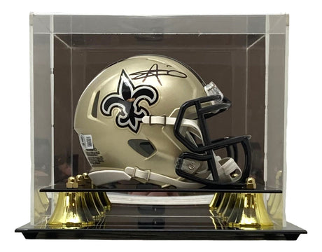 The Sports Integrity Alvin Kamara Signed New Orleans Saints Mini Speed Helmet, featuring a gold design with black facemask and team logo, is displayed in a clear case and mounted on a black and gold stand.