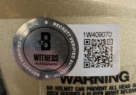A close-up of a Beckett Witness Autograph sticker with QR code and serial number 1W409070 on the Alvin Kamara Signed New Orleans Saints Mini Speed Helmet from Sports Integrity, with part of a warning label visible in the background.