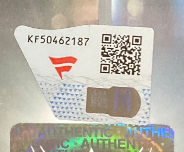 A close-up of a Sports Integrity security label for the Austin Theory Signed Framed 8x10 WWE Photo Fanatics, featuring code KF50462187, a red logo, QR code, and a holographic sticker with "AUTHENTIC" partially visible.