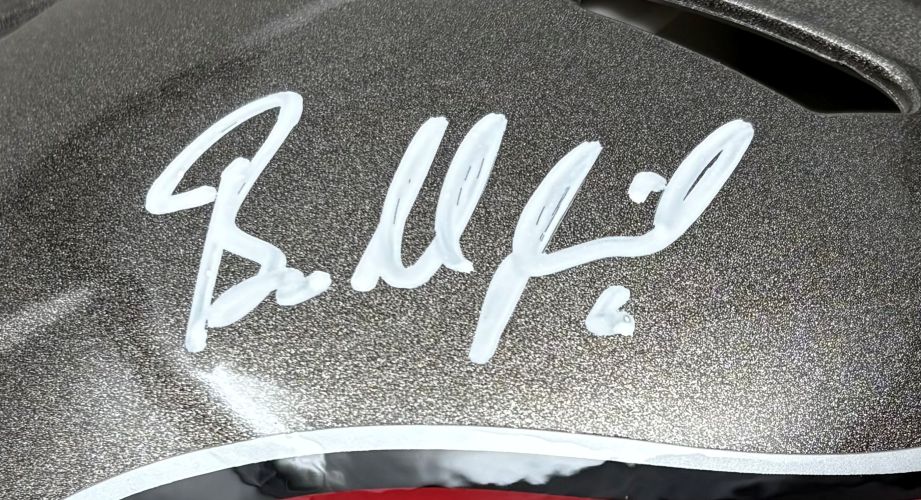 A close-up of the Baker Mayfield Signed Tampa Bay Buccaneers Full Size Authentic Helmet by Sports Integrity shows his white marker signature on a metallic gray surface, with black, white, and a hint of red visible at the helmet's edge.