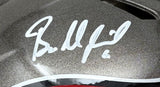 A close-up of the Baker Mayfield Signed Tampa Bay Buccaneers Full Size Authentic Helmet by Sports Integrity shows his white marker signature on a metallic gray surface, with black, white, and a hint of red visible at the helmet's edge.