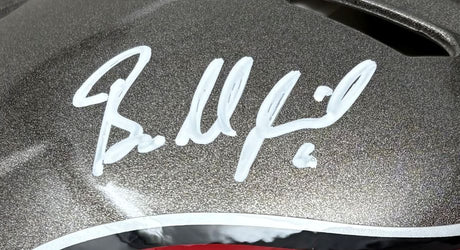 A close-up of the Baker Mayfield Signed Tampa Bay Buccaneers Full Size Authentic Helmet by Sports Integrity shows his white marker signature on a metallic gray surface, with black, white, and a hint of red visible at the helmet's edge.