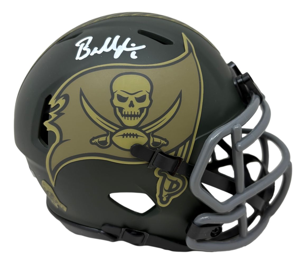 The Sports Integrity Baker Mayfield Signed Buccaneers Salute To Service Mini Speed Helmet features a matte black finish, gold flag logo with skull and crossed swords, silver facemask, and Baker Mayfield's autograph on top left.