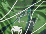 A close-up of Belal Muhammad Signed Custom Green MMA Fight Trunks by Sports Integrity features abstract green patterns, white lines, a white snake logo, and a bold silver autograph across the fabric.