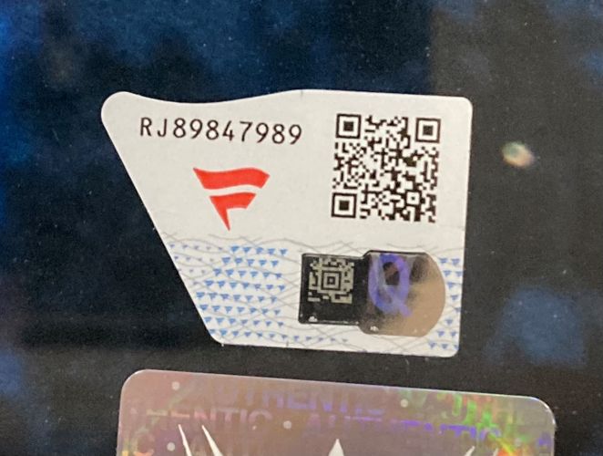 A close-up of a sticker with the code RJ89847989, red logo, QR code, and unique details on a reflective background from the Charlotte Flair Signed Framed 16x20 WWE 2025 Royal Rumble Photo by Sports Integrity.