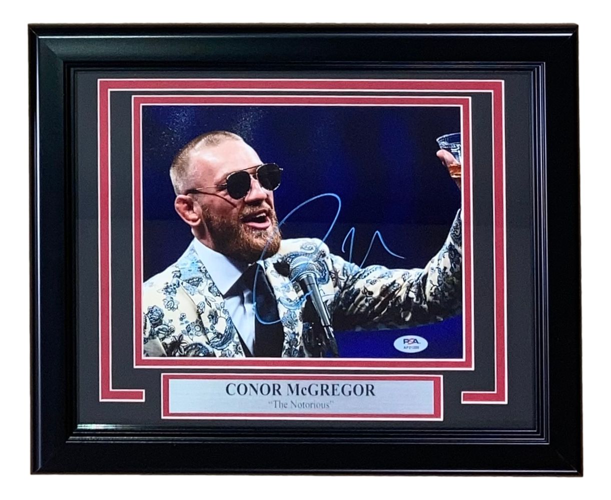 Sports Integrity Conor McGregor Signed Framed 8x10 UFC Photo (with PSA/DNA Hologram) featuring McGregor in sunglasses and a patterned suit, raising a glass and speaking; includes “CONOR McGREGOR ‘The Notorious.’” plaque.