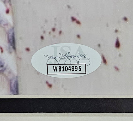 A close-up of an oval JSA sticker marked with code WB104895 appears on a Creed Bratton Signed Framed 11x14 The Office Creed Bloody Shirt Photo, authenticated by Sports Integrity, against a light background with blurred spots.