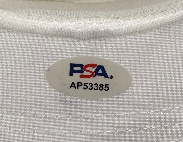 A close-up of the Sports Integrity John Daly Signed White Daly Adjustable Hat's fabric label, featuring an oval PSA/DNA sticker in red and blue with code AP53385.