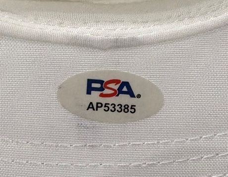 A close-up of the Sports Integrity John Daly Signed White Daly Adjustable Hat's fabric label, featuring an oval PSA/DNA sticker in red and blue with code AP53385.
