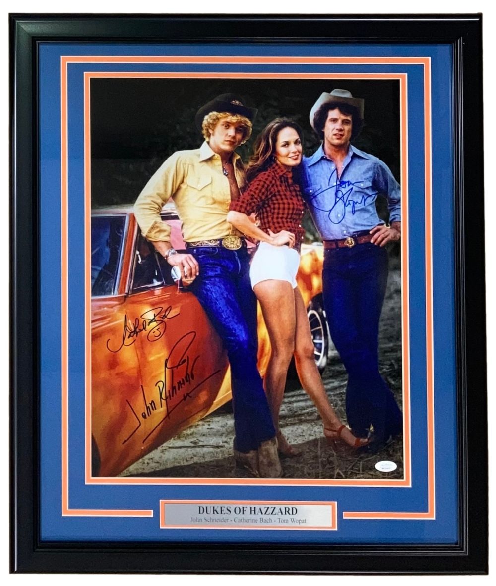 Sports Integrity Schneider, Wopat, Bach signed and framed 16x20 "Dukes of Hazzard" photo (JSA hologram), showing two men in jeans and a woman in shorts standing before an orange car; actors’ names on plaque below.