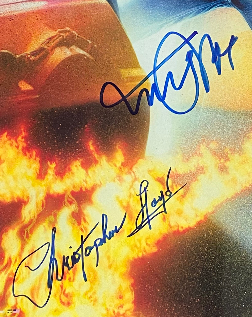 A close-up of blue Michael J Fox and Christopher Lloyd autographs on a fiery BTTF poster background, showing flames and a partial car tire. Sold by Sports Integrity, this signed framed piece includes PSA+JSA hologram authentication.