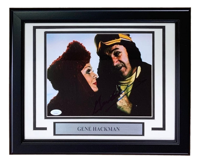 Sports Integrity's Gene Hackman Signed Framed 8x10 Superman II Photo features two costumed characters, one in a red hood and one in a yellow and brown headpiece. Includes a plaque with Gene Hackman's name. Authenticated by JSA.