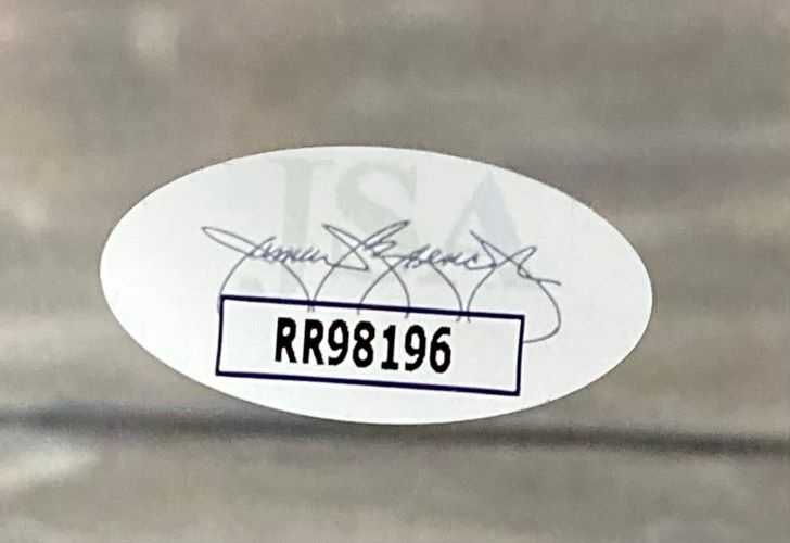 An oval JSA hologram with a faint signature and bold code RR98196 is affixed to the Giannis Antetokounmpo Signed Framed 11x14 Milwaukee Bucks Photo from Sports Integrity; the sticker sits inside a white box on a blurred gray background.