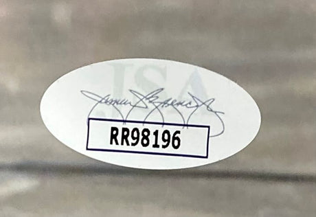 An oval JSA hologram with a faint signature and bold code RR98196 is affixed to the Giannis Antetokounmpo Signed Framed 11x14 Milwaukee Bucks Photo from Sports Integrity; the sticker sits inside a white box on a blurred gray background.