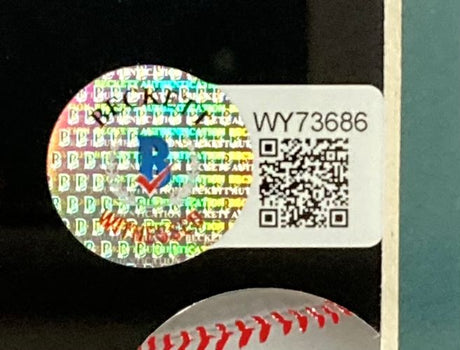 A round holographic sticker with repeating PB text, blue/red logo, and WY73686 beside a B&W QR code on a white label appears on the Sports Integrity Ken Griffey Jr Ichiro Suzuki Teal Signed Framed 16x20 Mariners Photo BAS. Part of a red baseball pattern shows underneath.