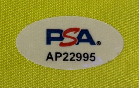 A white oval PSA/DNA hologram sticker (code AP22995) is attached to the Jordan Spieth Signed Framed The Memorial Tournament Golf Flag from Sports Integrity, shown on a textured yellow surface.