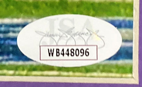 A white oval sticker with WB448096 in black appears on the Justin Tucker Signed Framed 16x20 Ravens Record Kick Photo by Sports Integrity, against a colorful blurred background. Faded text and a faint logo are also visible on the sticker.