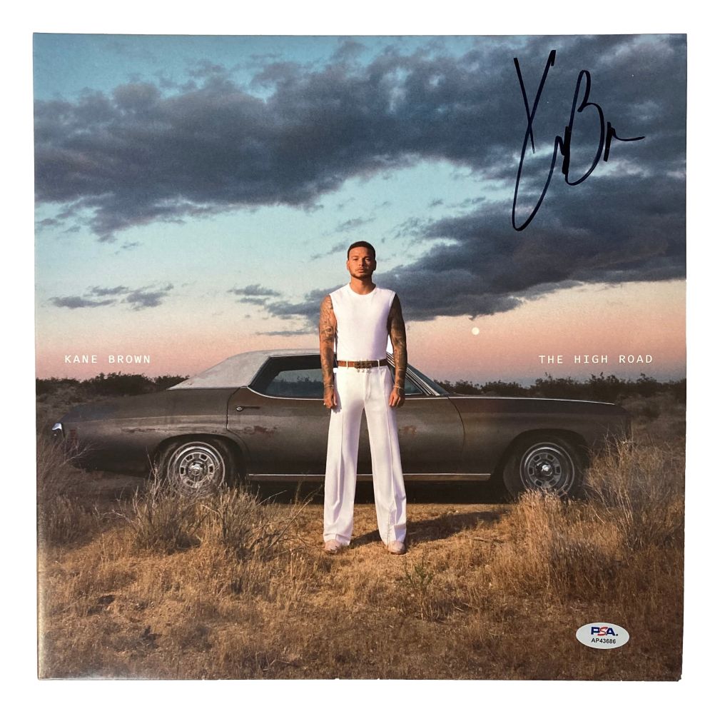 A man in a white sleeveless shirt and pants stands before a vintage car at sunset on the Kane Brown Signed The High Road Vinyl Record Cover by Sports Integrity, authenticated by PSA/DNA, with his signature above.