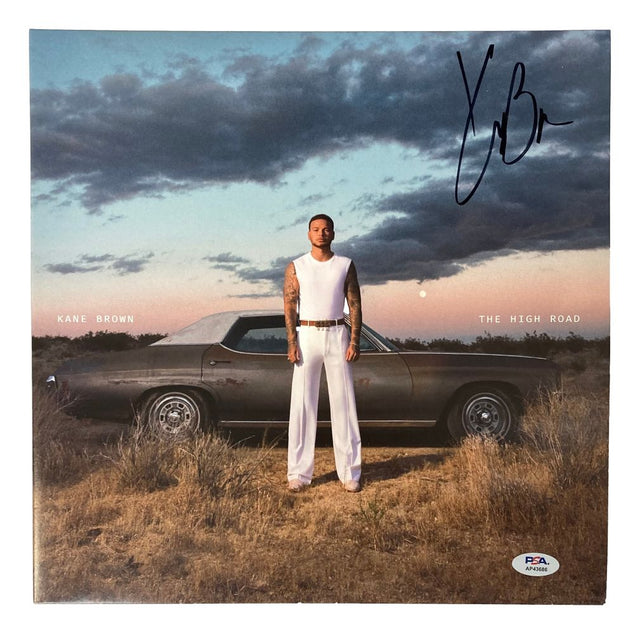 A man in a white sleeveless shirt and pants stands before a vintage car at sunset on the Kane Brown Signed The High Road Vinyl Record Cover by Sports Integrity, authenticated by PSA/DNA, with his signature above.