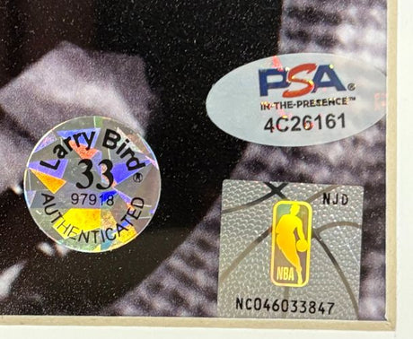 Close-up of three authentication stickers on a Sports Integrity Larry Bird Signed Framed 16x20 Celtics w/ Red Auerbach Photo: holographic Bird AUTHENTICATED, silver PSA 4C26161, and NBA logo NC046033847.