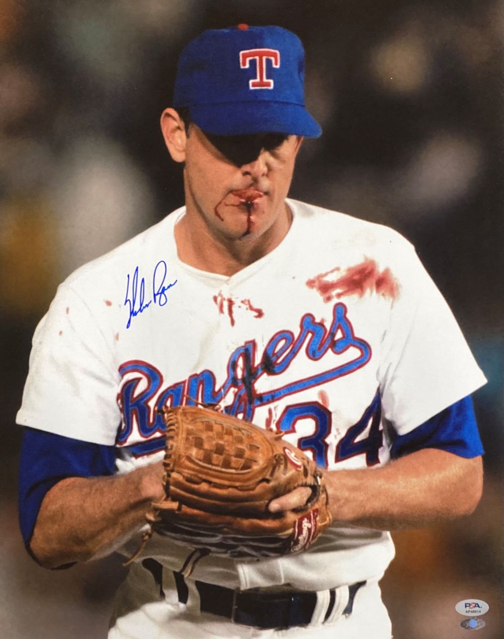 The Sports Integrity Nolan Ryan Signed 16x20 Texas Rangers Bloody Lip Photo (PSA/DNA) shows pitcher Nolan Ryan in a white #34 jersey with a blue cap, standing on the field with blood on his mouth and jersey during a game.