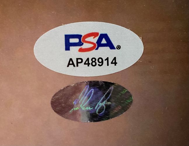 Two oval stickers are on a brown surface: the top is white with PSA AP48914 in blue, red, and black; the bottom is metallic with indistinct writing. This relates to the Sports Integrity Nolan Ryan Signed 16x20 Texas Rangers Bloody Lip Photo PSA/DNA.