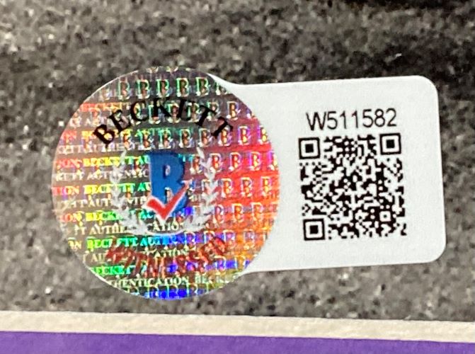 A holographic Beckett sticker with QR code and serial W511582 is attached to the Ray Lewis Signed Framed 8x10 Baltimore Ravens Flag Photo by Sports Integrity, featuring Beckett logo and multicolored security text on a dark textured surface.