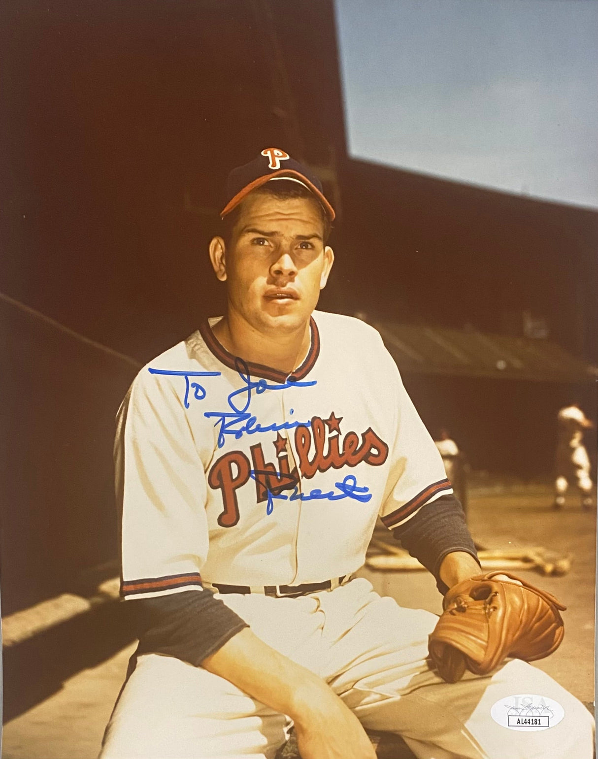 The Sports Integrity Robin Roberts Signed 8x10 Philadelphia Phillies Photo (JSA AL44181) features Roberts in uniform holding a glove in the dugout, with a bat and another player visible in the background. Signed in blue ink.