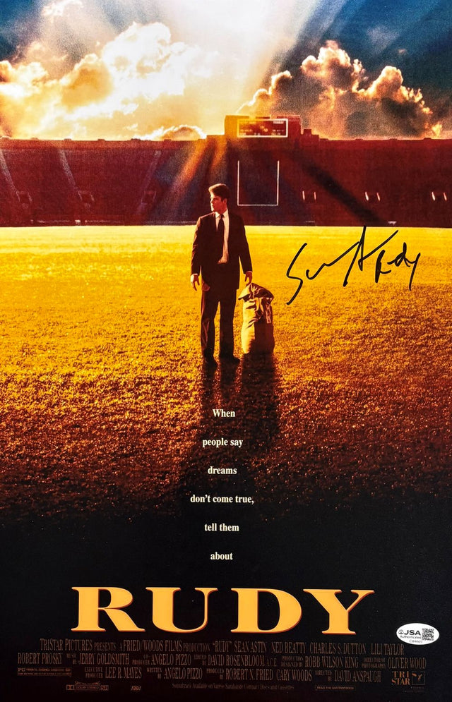 A young man stands on a sunlit football field, holding a sports bag as he faces empty stadium seats. Text: "When people say dreams don't come true, tell them about Rudy." Sports Integrity Sean Astin Signed 11x17 Rudy Photo JSA.